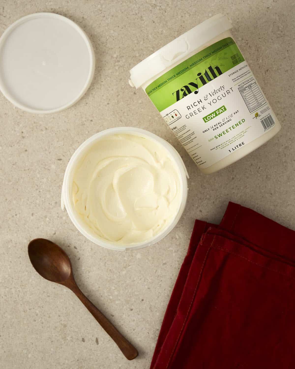 An open tub of Zayith Low-Fat Sweetened Greek Yogurt beside the 1 litre tub, with a wooden spoon